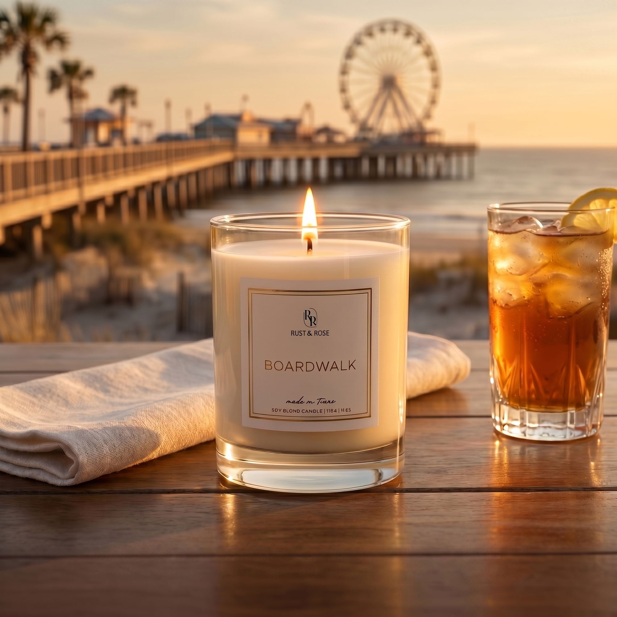Candle labeled 'Boardwalk' by Rust & Rose on a beach setting with a Ferris wheel in the background.